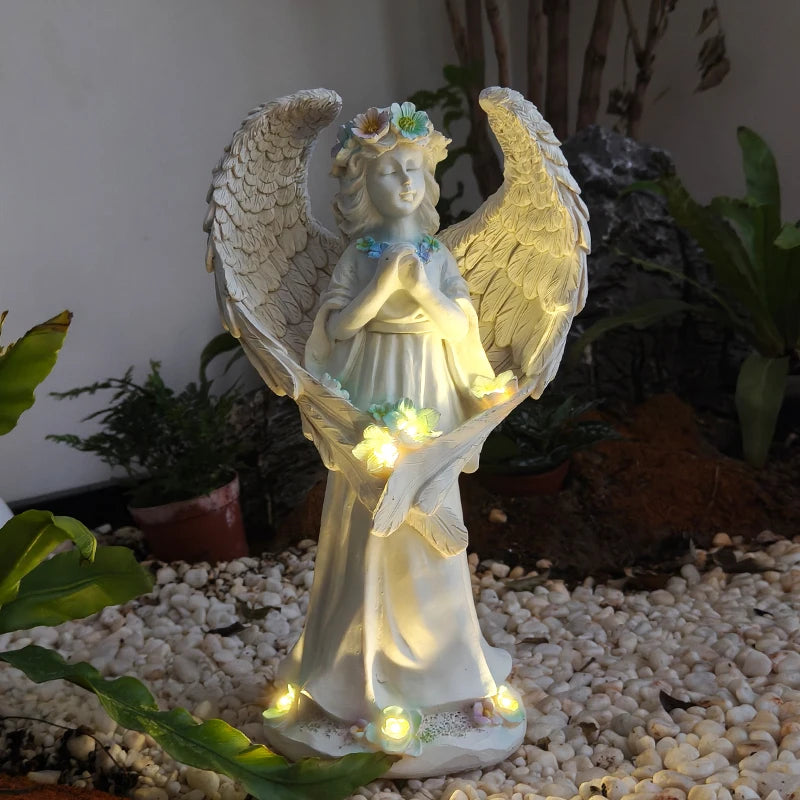 An angel garden statue with outstretched wings, a floral crown, and warm white LED lights glowing around the base and near the hands, displayed outdoors on a bed of white pebbles with plants in the background.