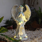An angel garden statue with outstretched wings, a floral crown, and warm white LED lights glowing around the base and near the hands, displayed outdoors on a bed of white pebbles with plants in the background.